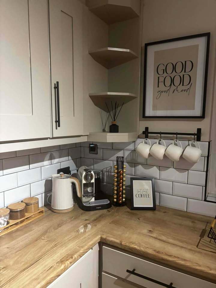 A kitchen corner with light wood countertops, white subway tile backsplash, and cream cabinets.  Coffee maker, kettle, mugs, and coffee pods are visible.  Wall art reads "Good Food Good Mood".