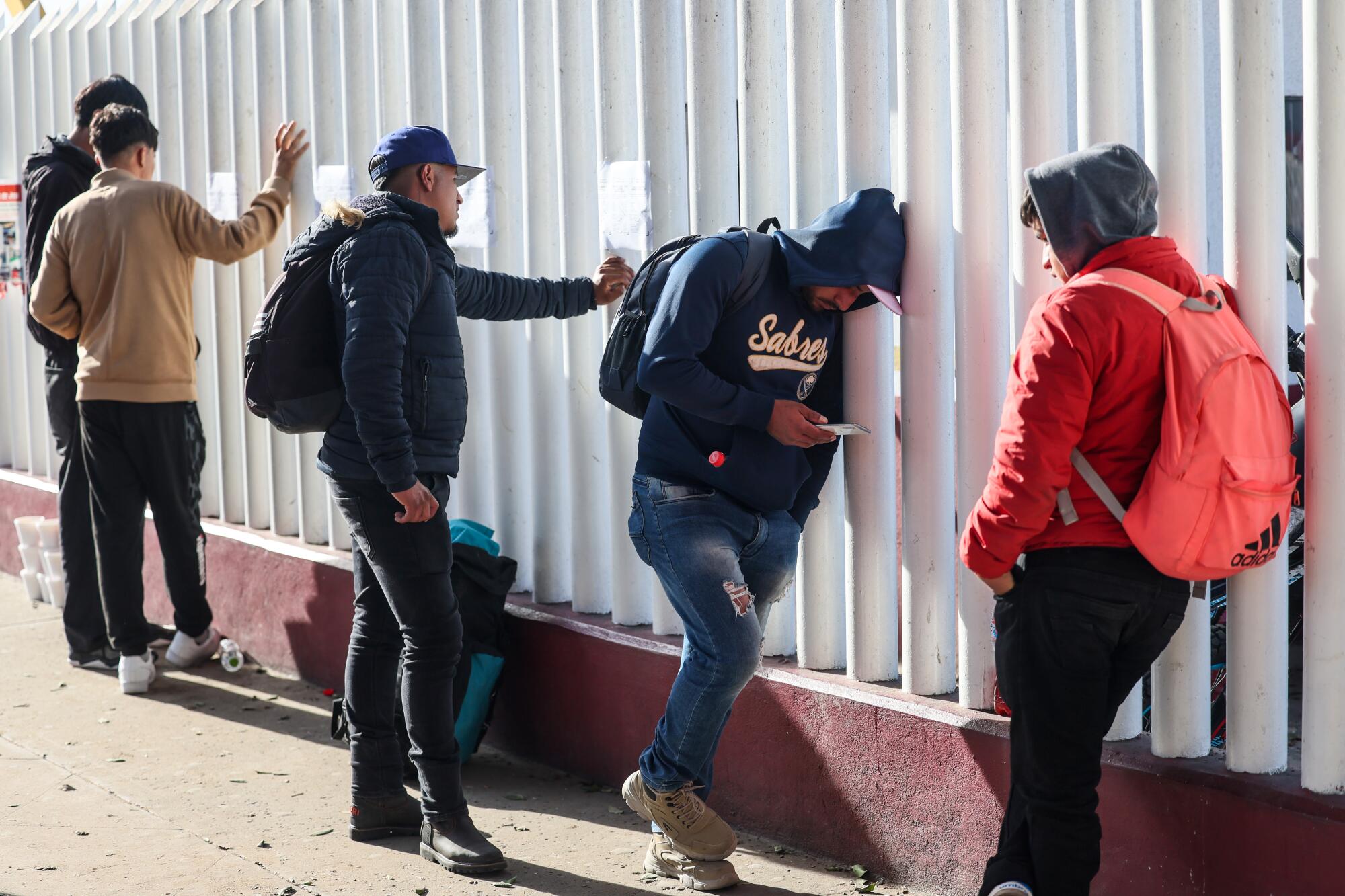 Asylum seekers lean against a white fence outside a customs office in Tijuana.