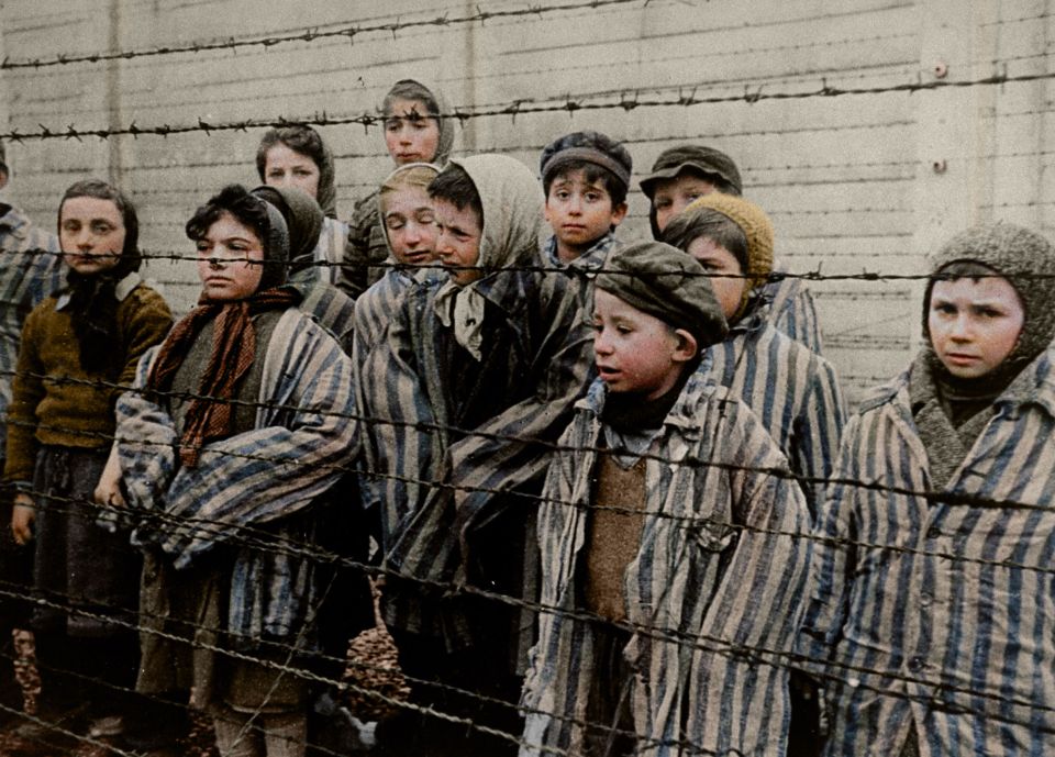 Colorized photo of Jewish children at Auschwitz behind barbed wire.