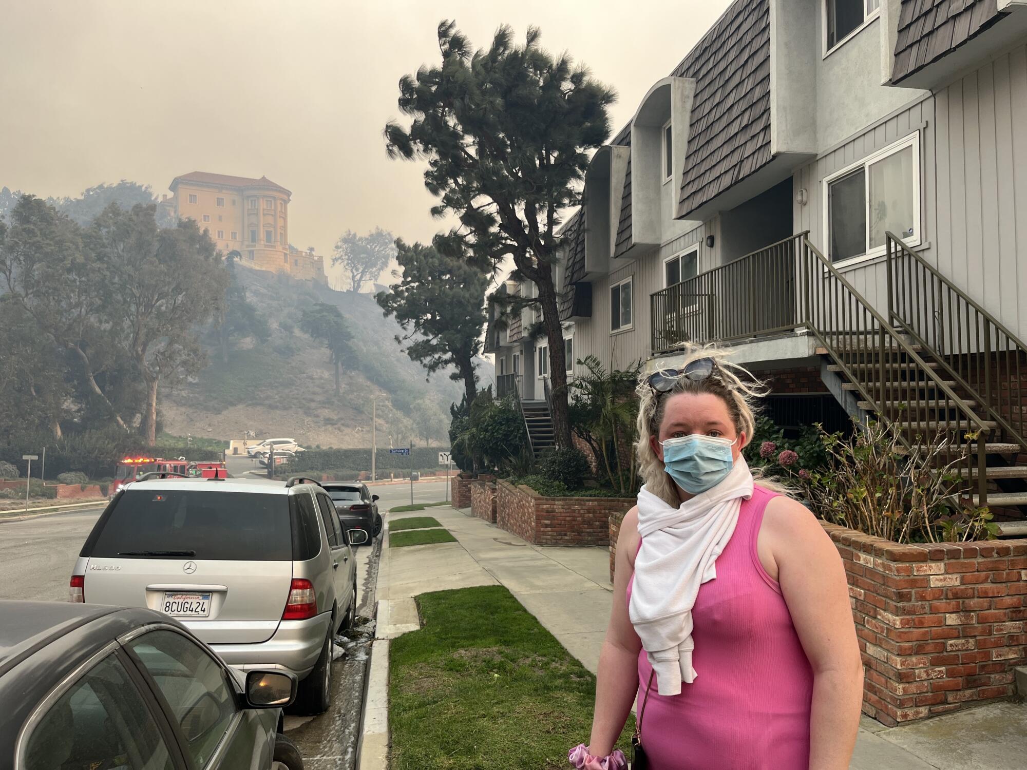 A woman in a mask stands outside a building with smoke in the background