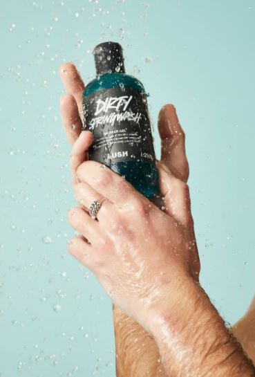 Hands holding a bottle of Lush Dirty Springwash shower gel under running water.