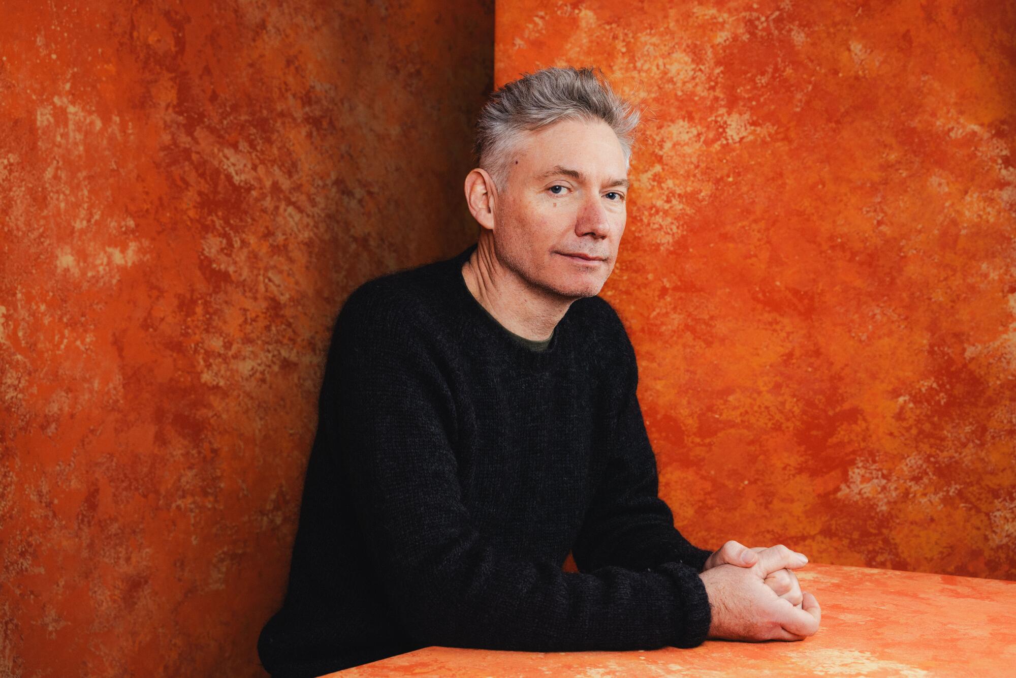 A gray-haired man in a black sweater sits at an orange table before an orange backdrop