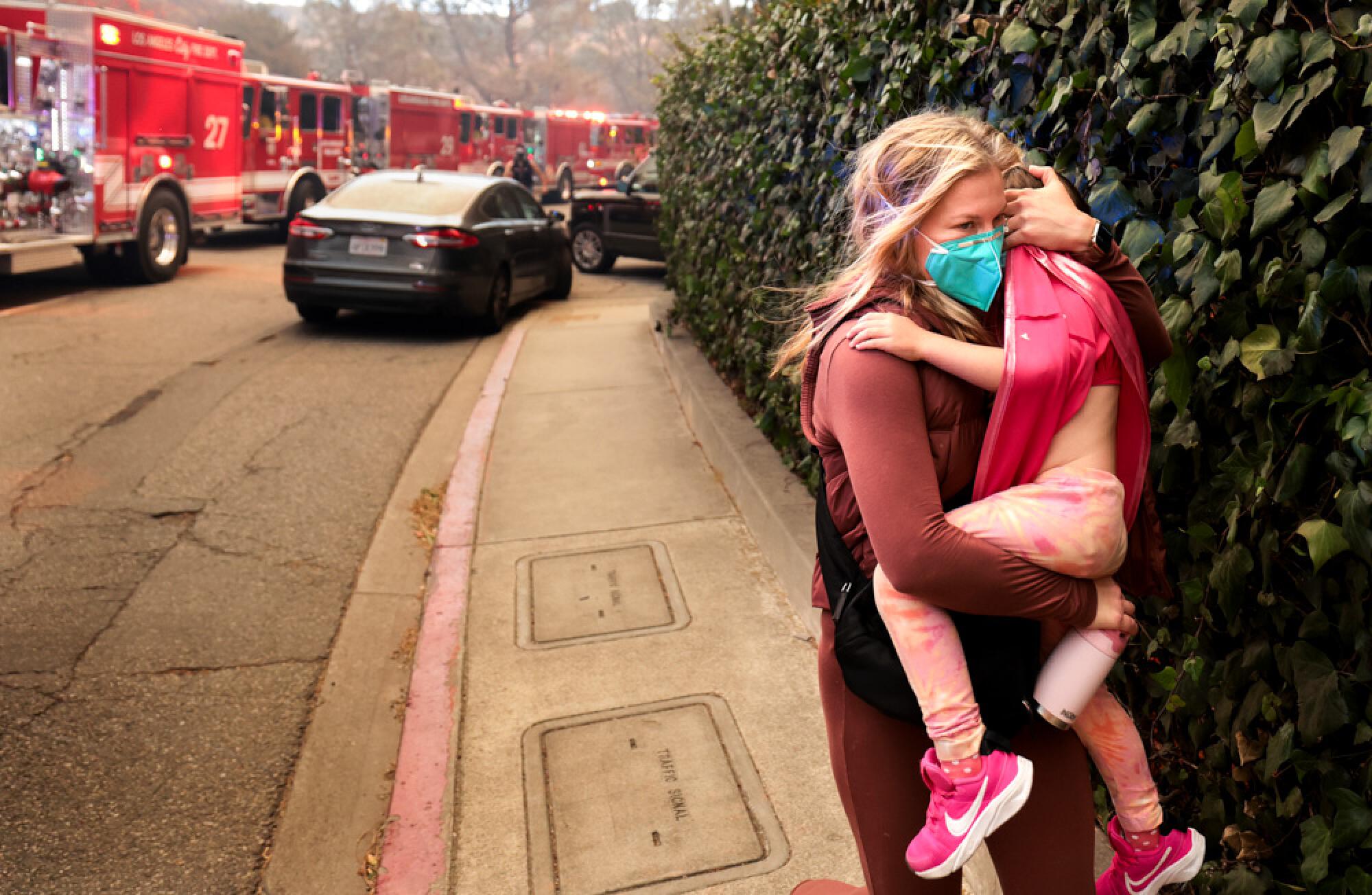 A mother carries her child as they move down a sidewalk away from a line of firetrucks. 