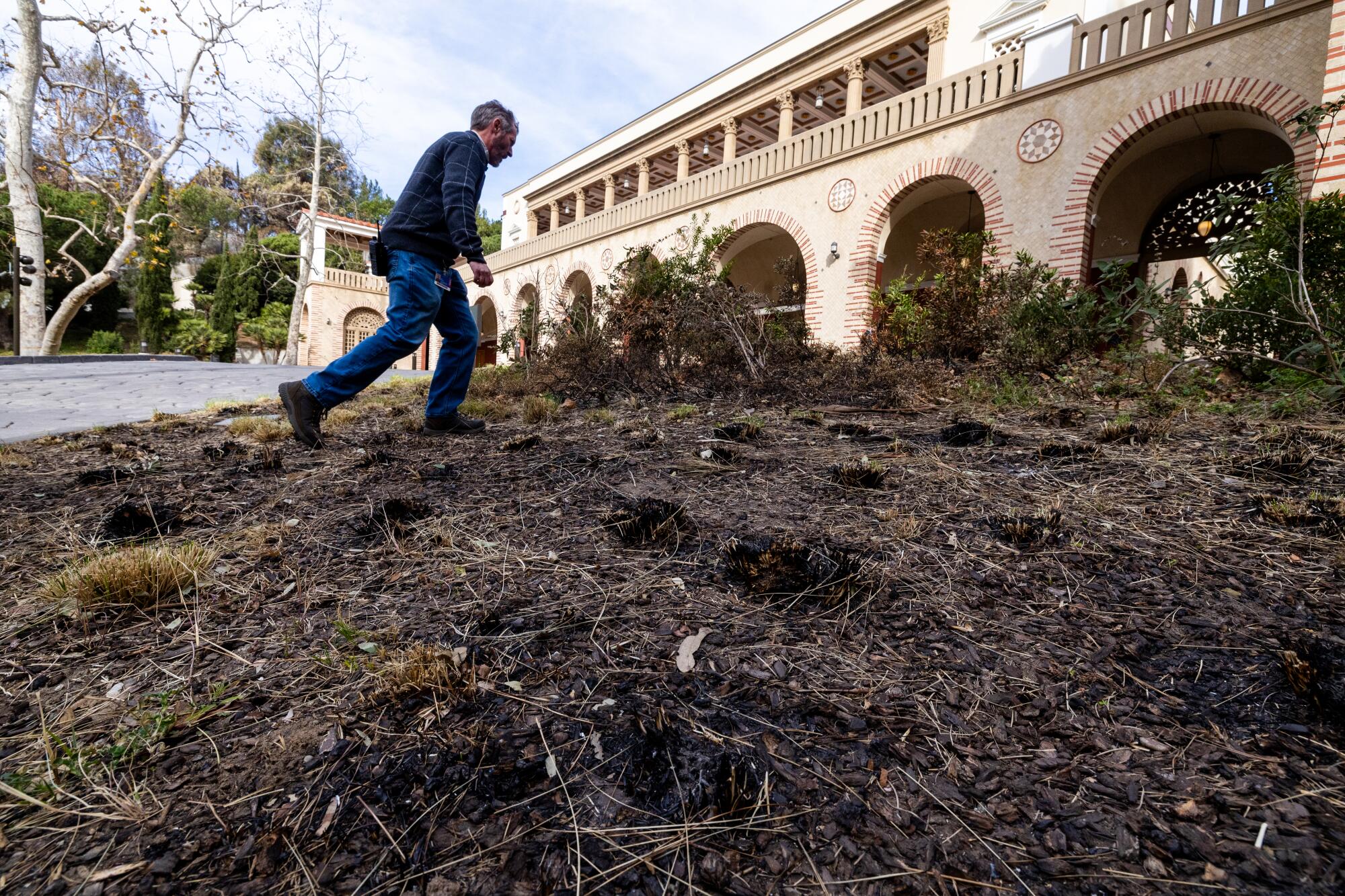 A man strides across burned landscaping toward a structure with numerous archways.