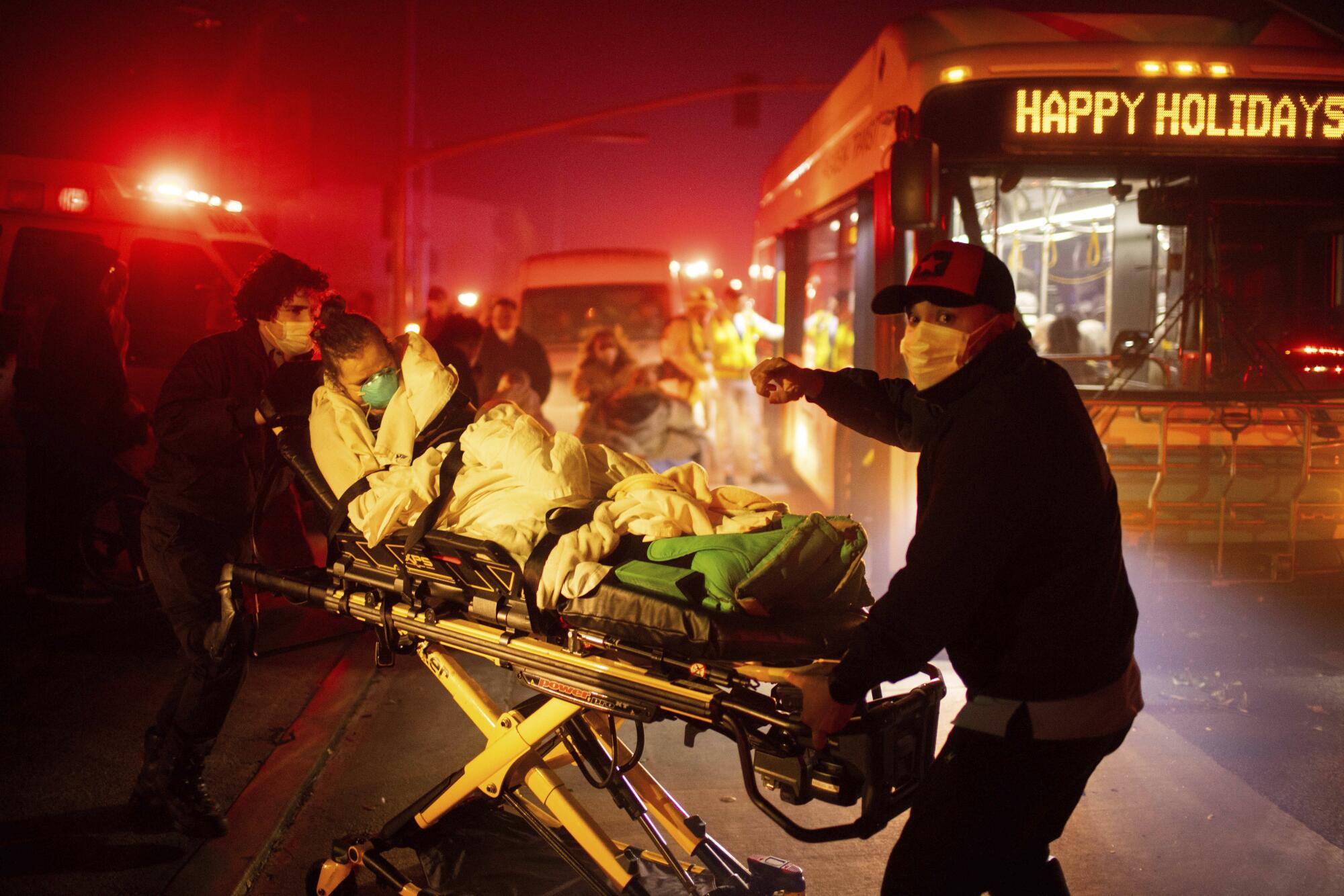 Two people with face masks prepare to load a patient on a gurney onto a bus.