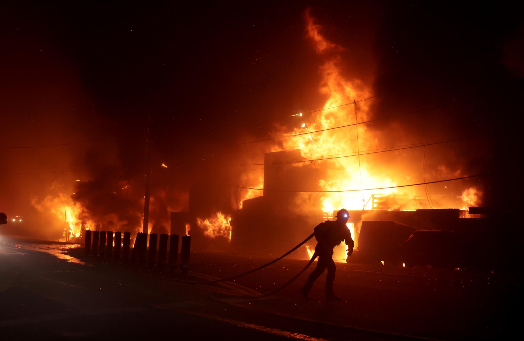 A firefighter pulling a hose is silhouetted by the light of a flaming structure.