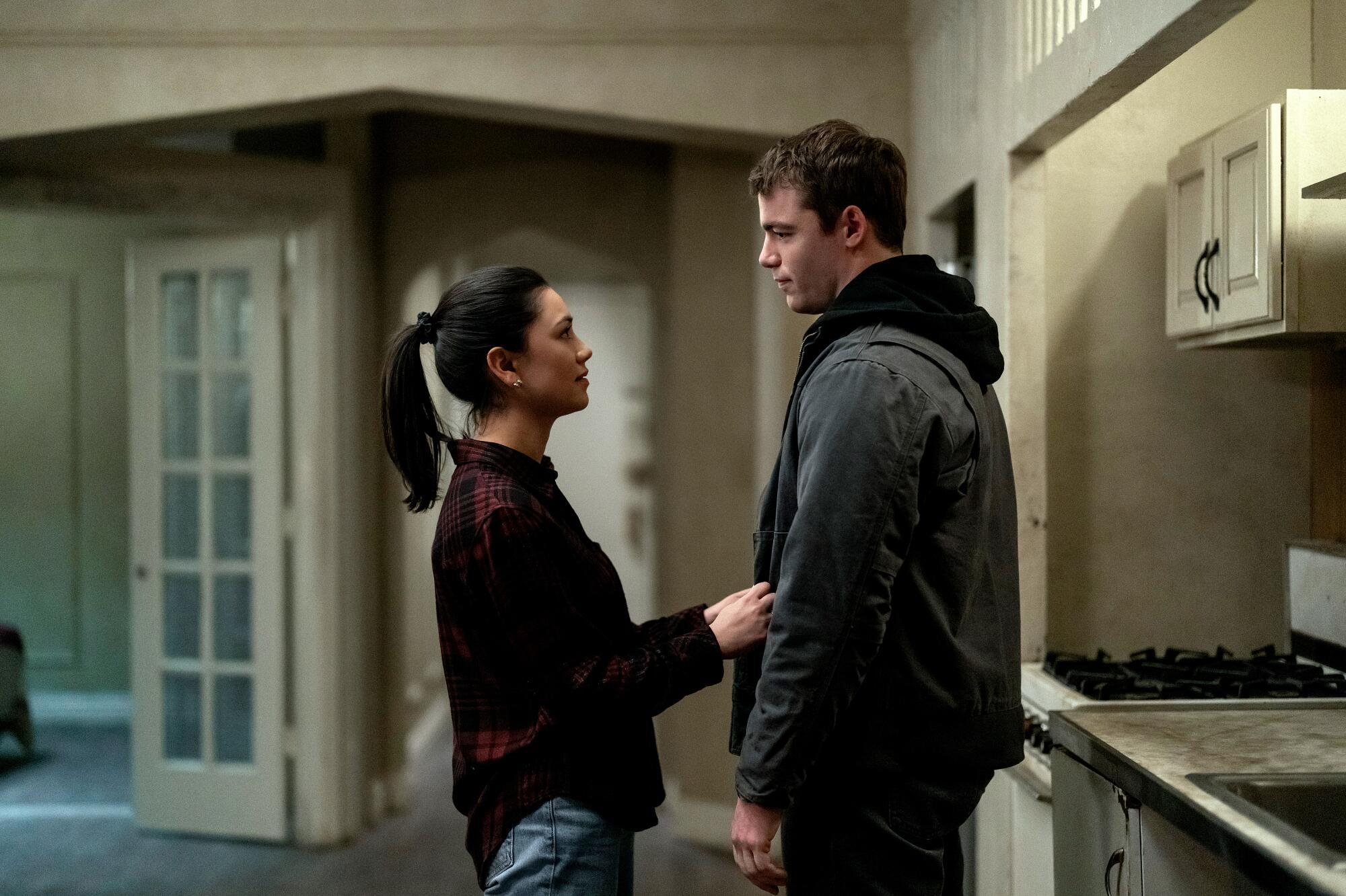 A woman and a man stand facing each other in a kitchen. She's holding onto his open jacket.