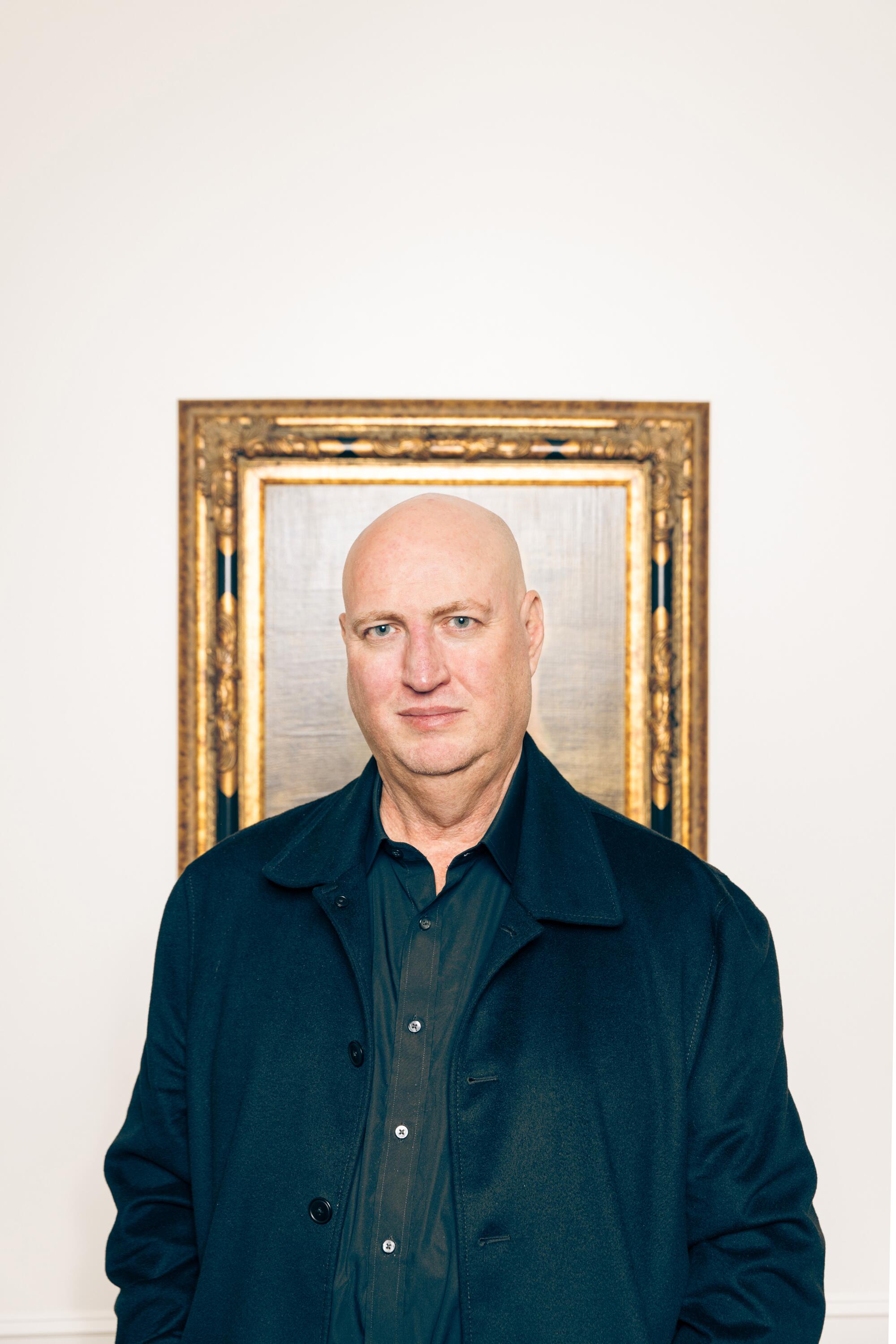 A bald man stands in front of a gold picture frame