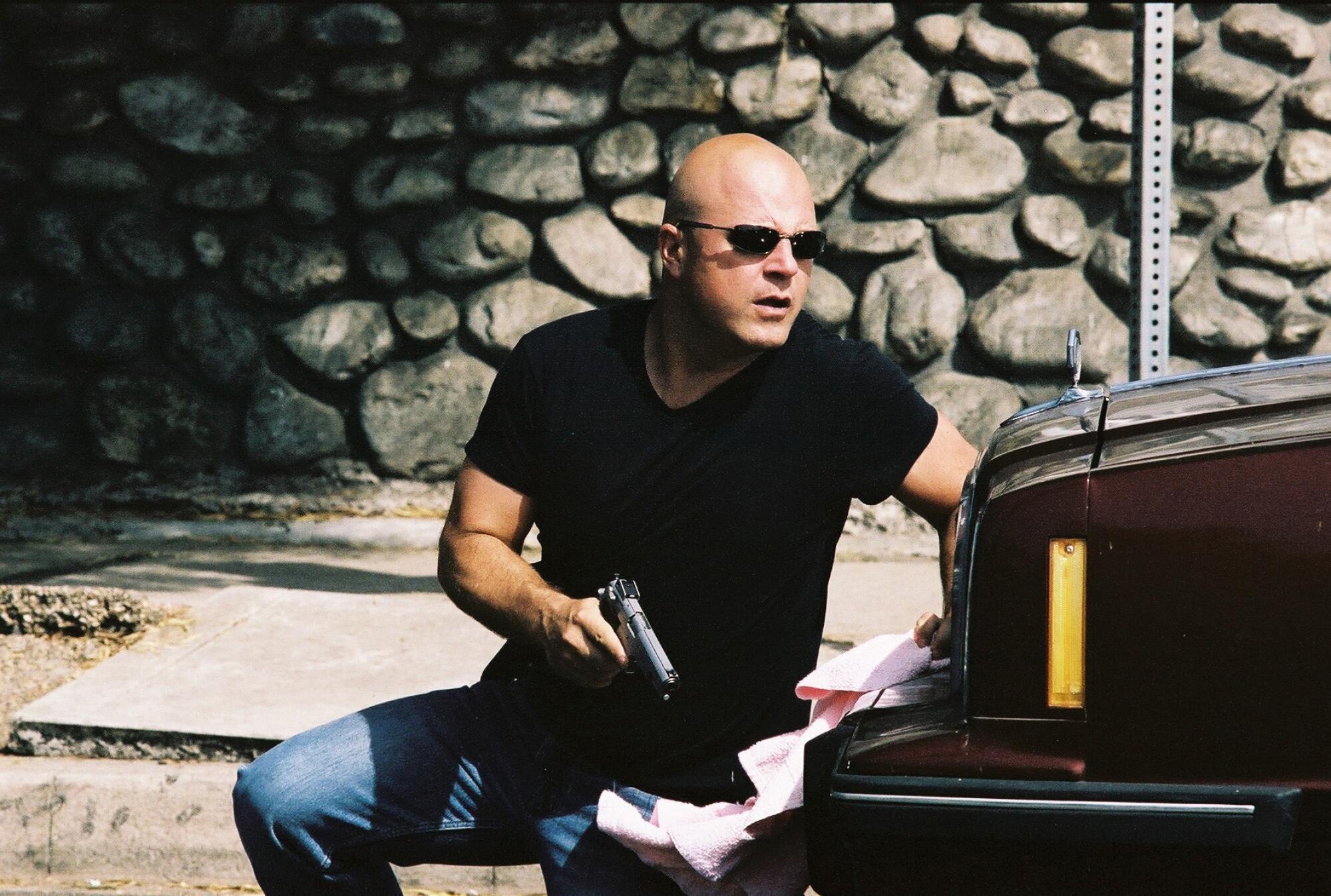 A bald man wearing sunglasses and a black T-shirt holding a gun and crouching behind a car