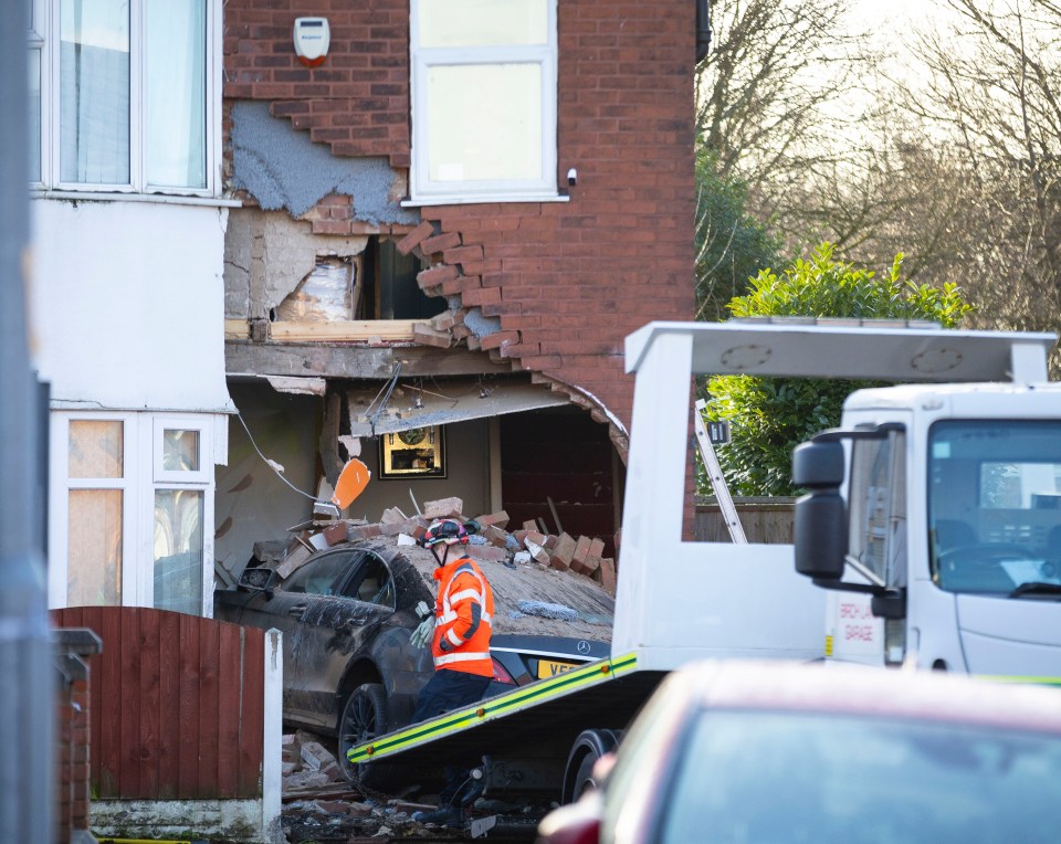 A car crashed into a house, causing significant damage to the building's brickwork.  A worker is loading the car onto a tow truck.