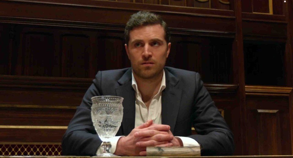 Man in suit sitting at a table, hands clasped.