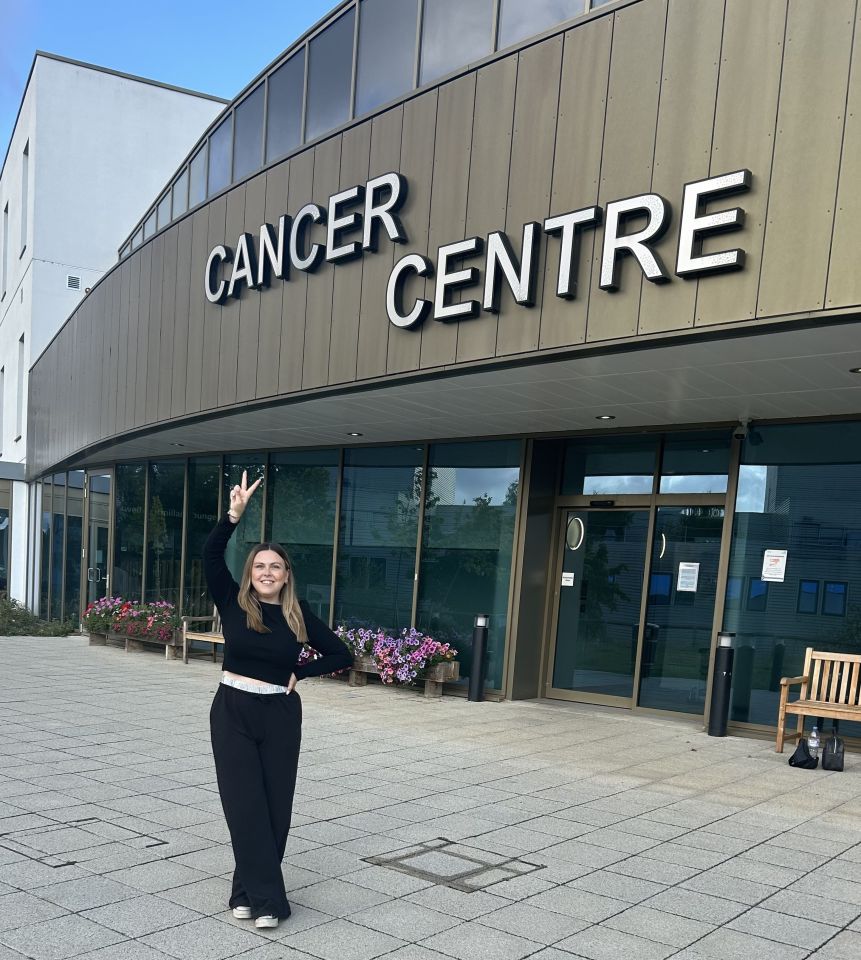 Woman standing in front of a cancer center, giving a peace sign.