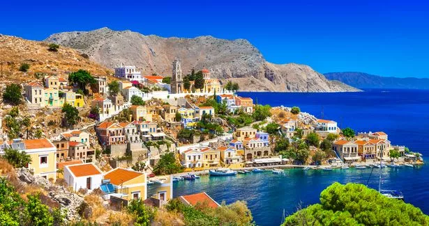 An aerial shot of Symi Island during the day, showing the islands colourful stone houses and clear blue water.