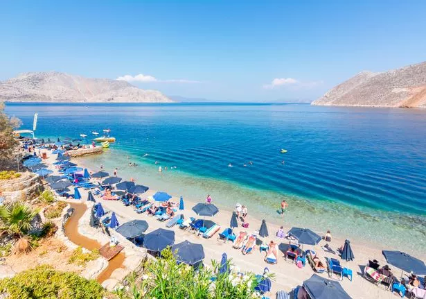 An aerial shot showing beachgoers on Nos Beach on Symi island, featuring rows of sunloungers and umbrellas