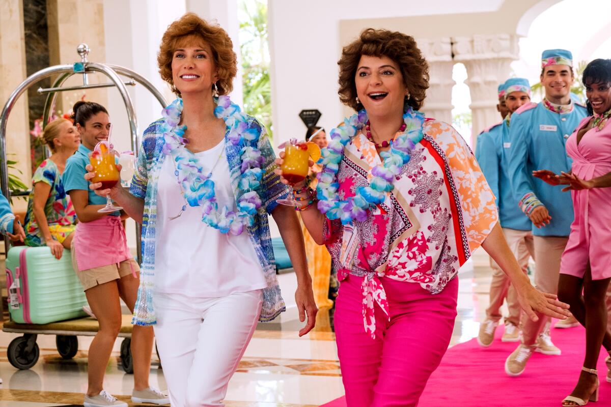 Two women wearing leis walk through a hotel lobby in the 2021 movie "Barb & Star Go to Vista Del Mar."