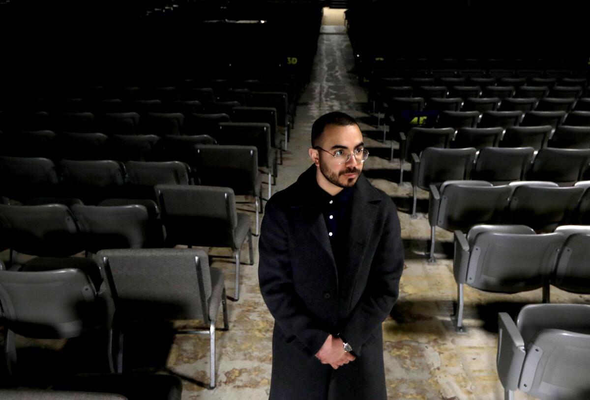 A man with dark hair and beard, wearing glasses and dark clothing, stands with hands clasped in a room with rows of chairs
