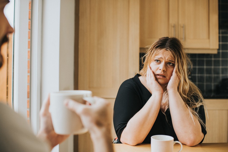 An anxious woman sits at a kitchen table with a man; she looks at him with a furrowed brow.