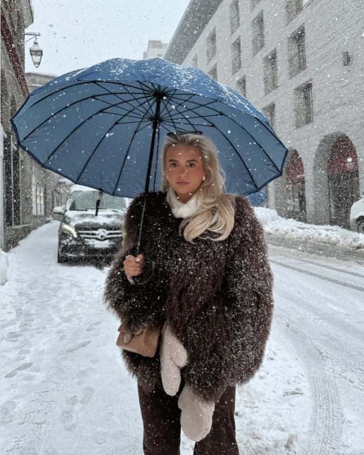 Woman in a brown fur coat under a blue umbrella in the snow.