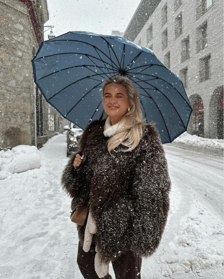 Woman in a brown fur coat under a blue umbrella in the snow.