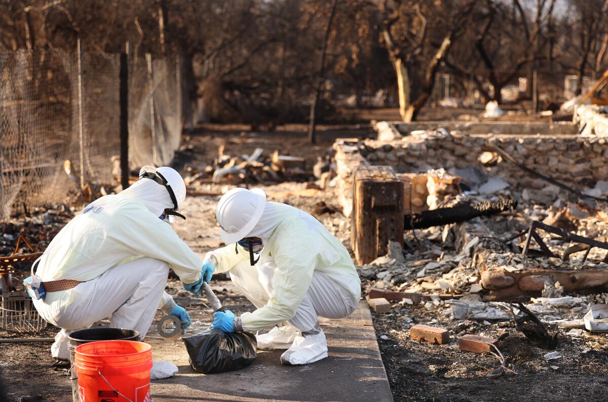 Two people in white protective gear, helmets and respirators seal hazardous items inside a plastic bag.