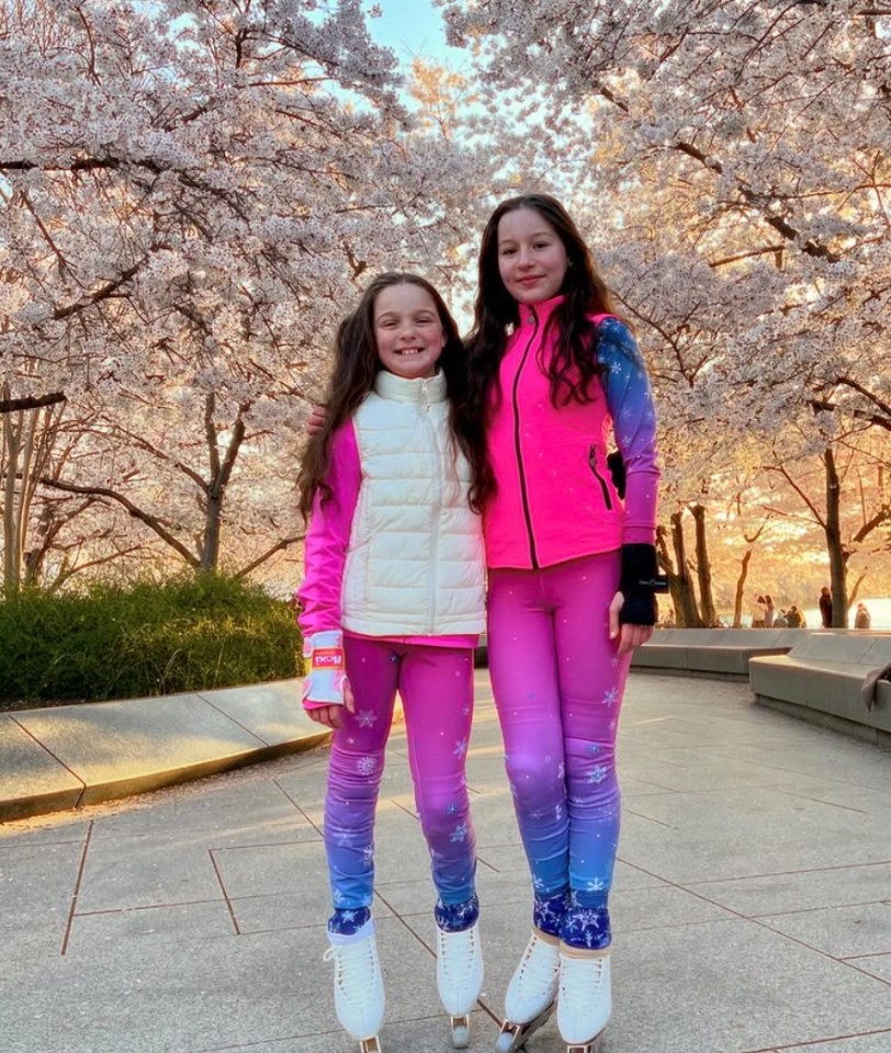 Two girls in ice skating outfits stand in front of cherry blossoms.