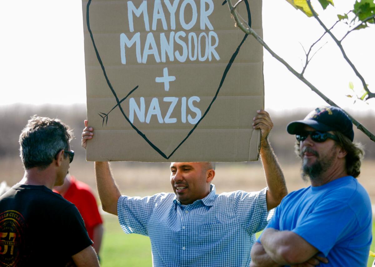 A man holds a sign that says "Mayor Mansoor + Nazis."