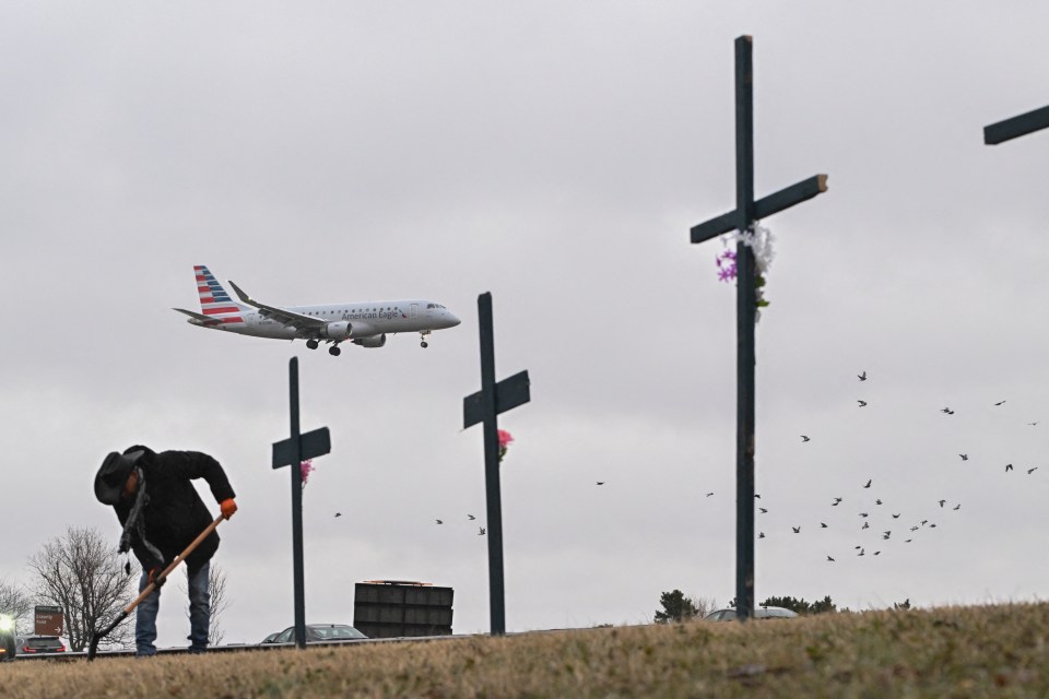 A person tends to a makeshift memorial of crosses near an airport, with a plane overhead.