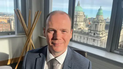 BBC Communities minister Gordon Lyons sits in front of a series of windows out of which the belfast skyline, including city hall, can be seen. He is balding, wearinhg a grey suit jacket , white shirt and grey tie. 