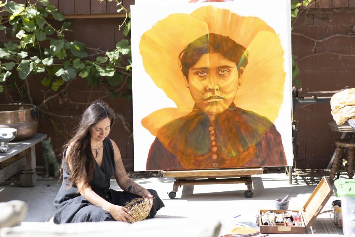 Artist Weshoyot Alvitre sits on the ground next to her painting of a Native American woman behind a California poppy