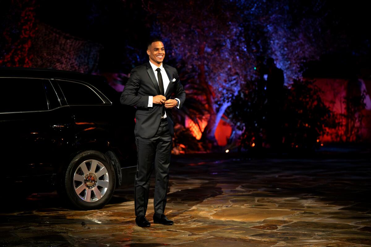 A man in a black suit and tie buttoning his jacket as he stands next to a limousine.