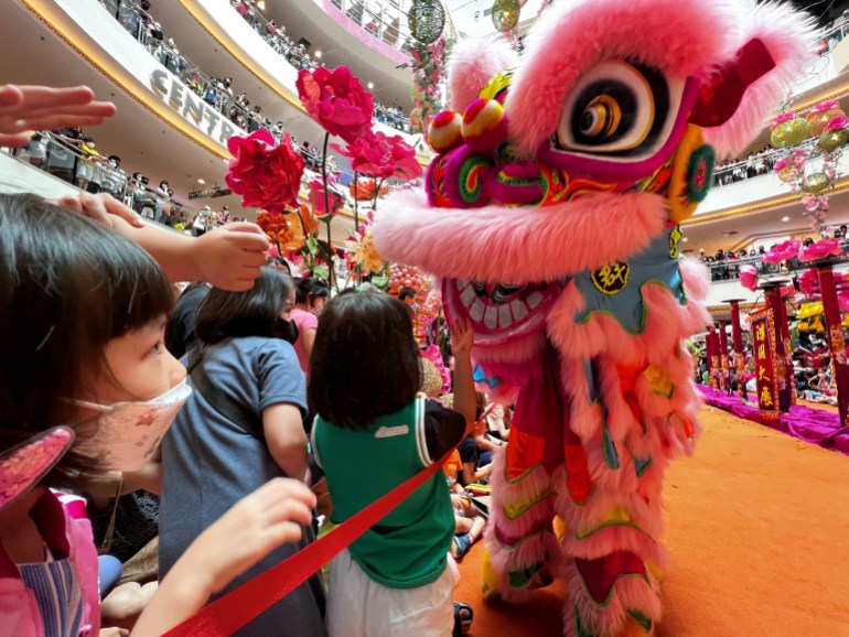 Children gather around a lion after a performance in Malaysia to get mandarin oranges. The lion has big eyes rimmed in pink fur . Its ears and top lip are also covered in pink fur. The children look excited. There are lots of people looking down on the concourse from higher levels.