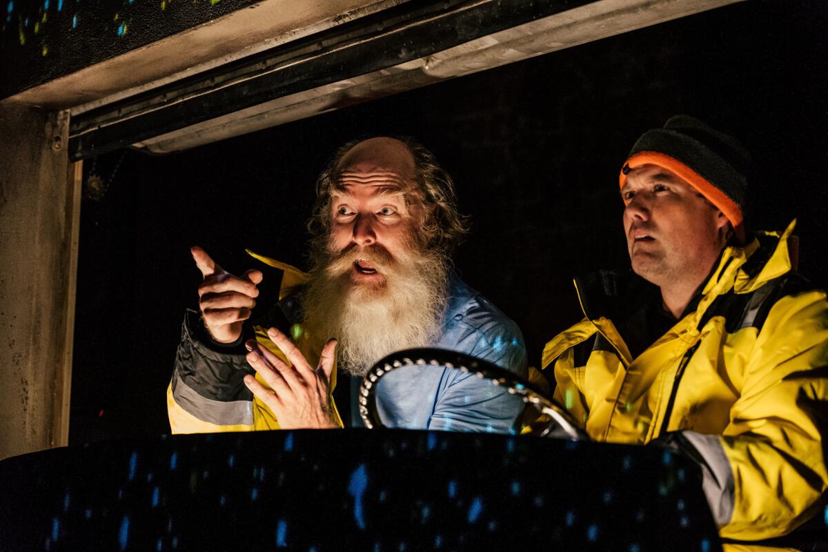 Onstage, two men in cold-weather gear, lit from below, appear to be in the cab of a truck.