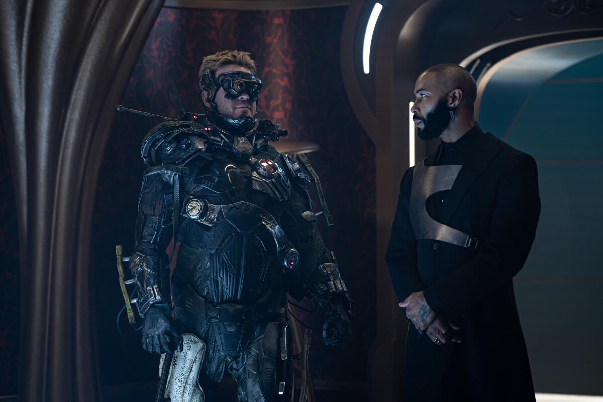 A man in an armored suit and goggles looks at a man in a black uniform.