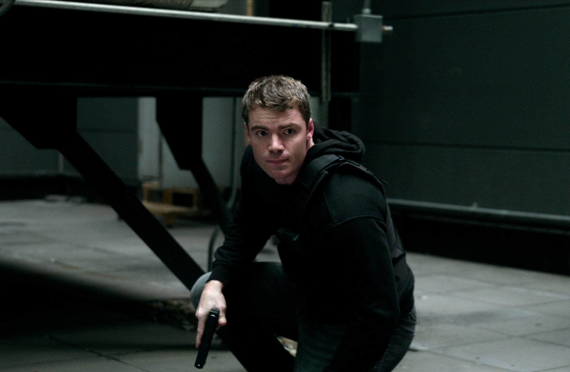 A man wearing a black hoodie and holding a gun is crouching to the ground