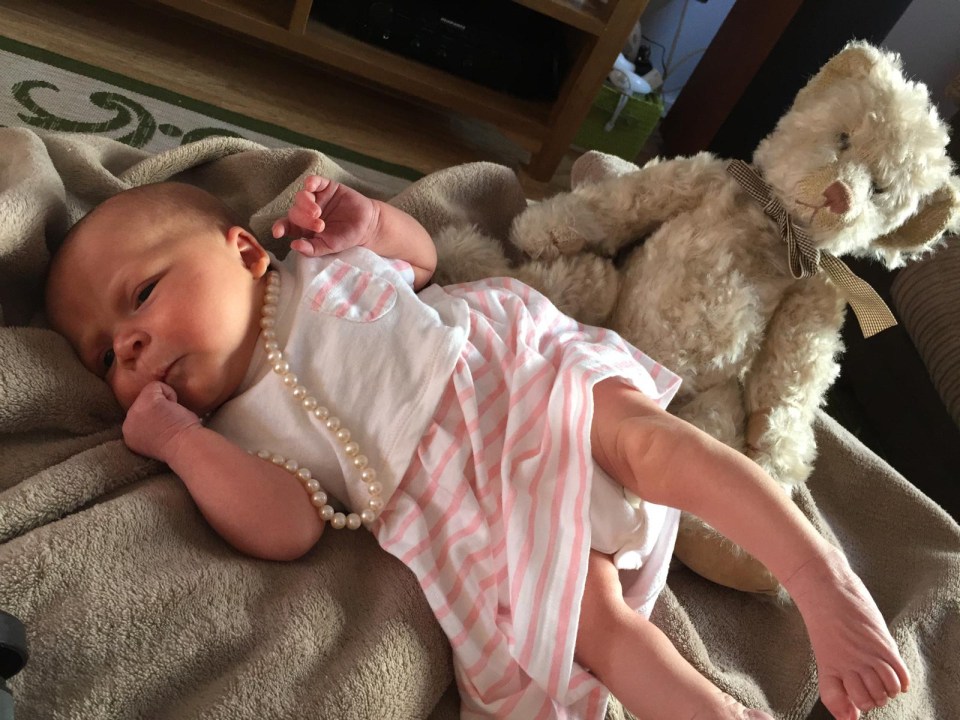 Baby girl wearing a pink and white striped dress and a pearl necklace, lying next to a teddy bear.