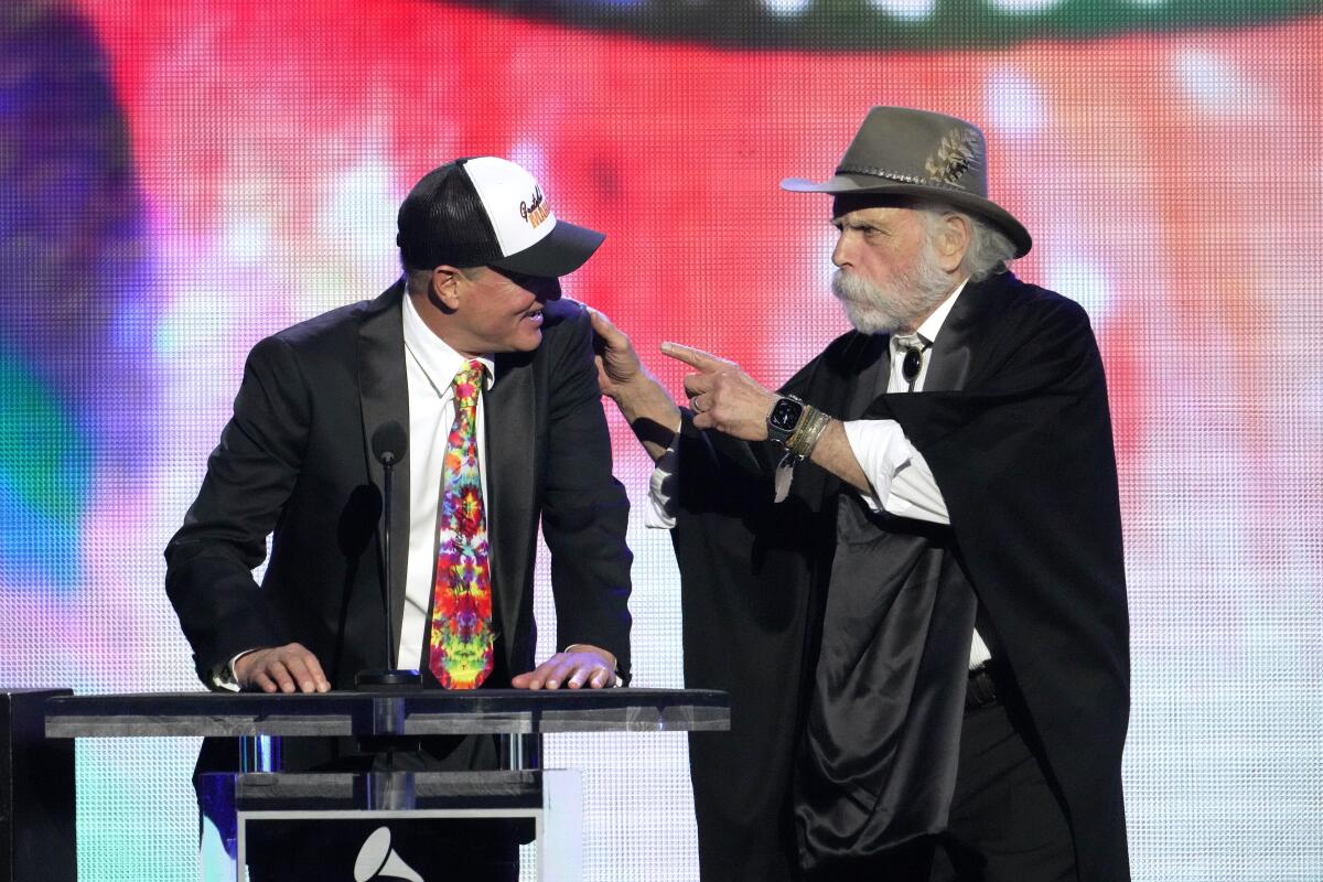 Woody Harrelson in a tie-dyed tie and black suit and Bob Weir in a black suit speak on stage while Weir points at Harrelson.