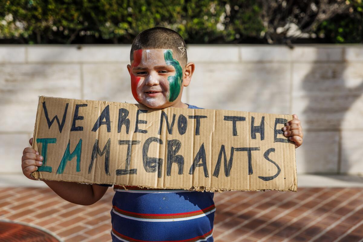 A child with a close-shaved head, green and red colors on his cheeks, holds a sign saying, "We are not the immigrants"