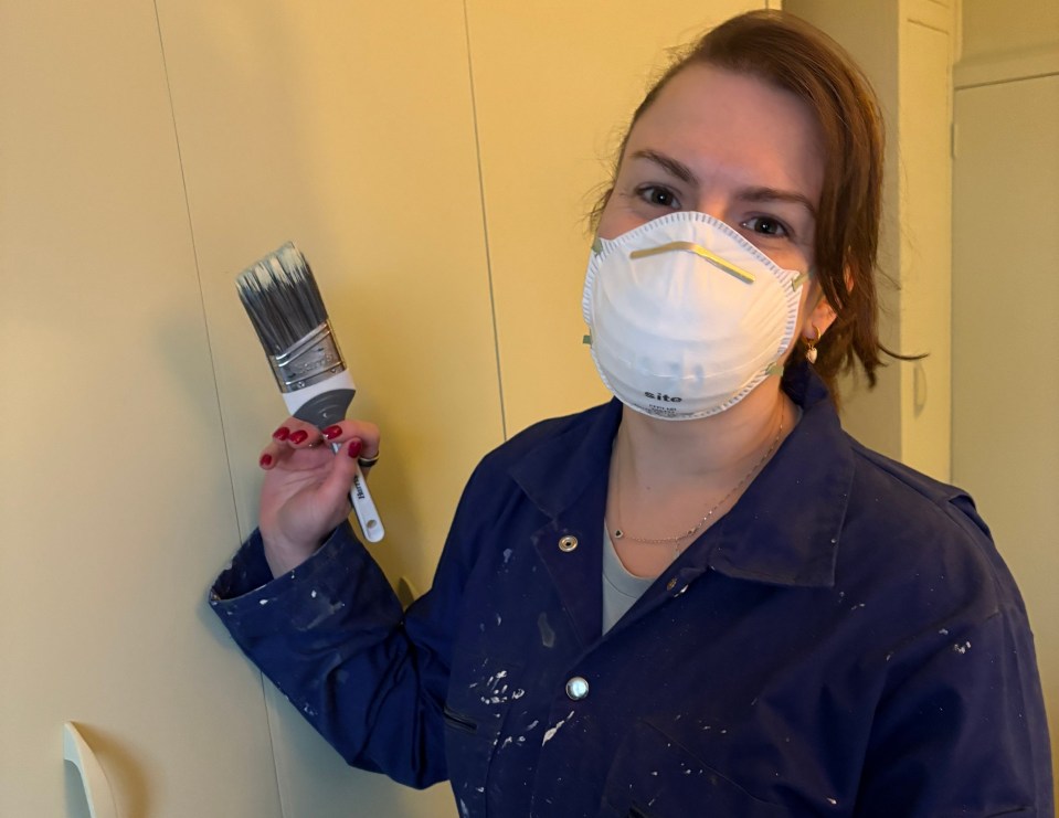 Woman in a paint-splattered jumpsuit and respirator mask holds a paintbrush.