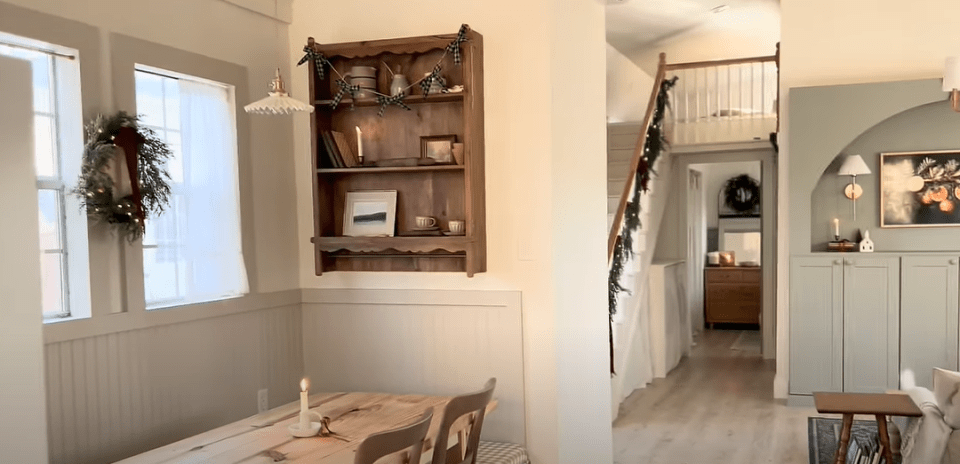 Interior view of a small home's dining area and hallway, showing a wooden shelf, dining table, and stairs leading to a loft.