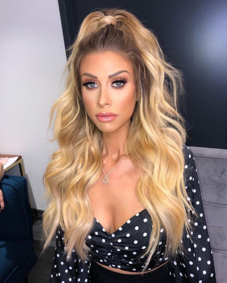 Woman with long blonde hair styled in loose waves and a half-updo, wearing a black polka dot top.