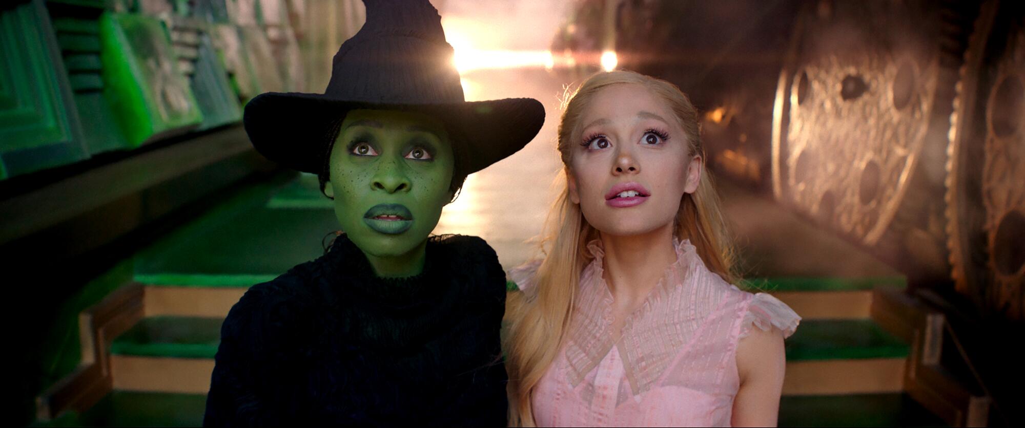 A woman in black with green skin stands with a woman in pink gazing up at the sky in "Wicked."