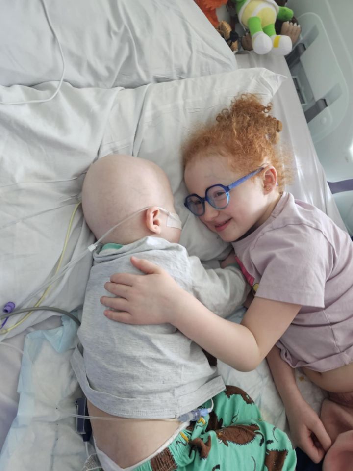 A young boy with stage four cancer lies in a hospital bed, hugged by his older sister.