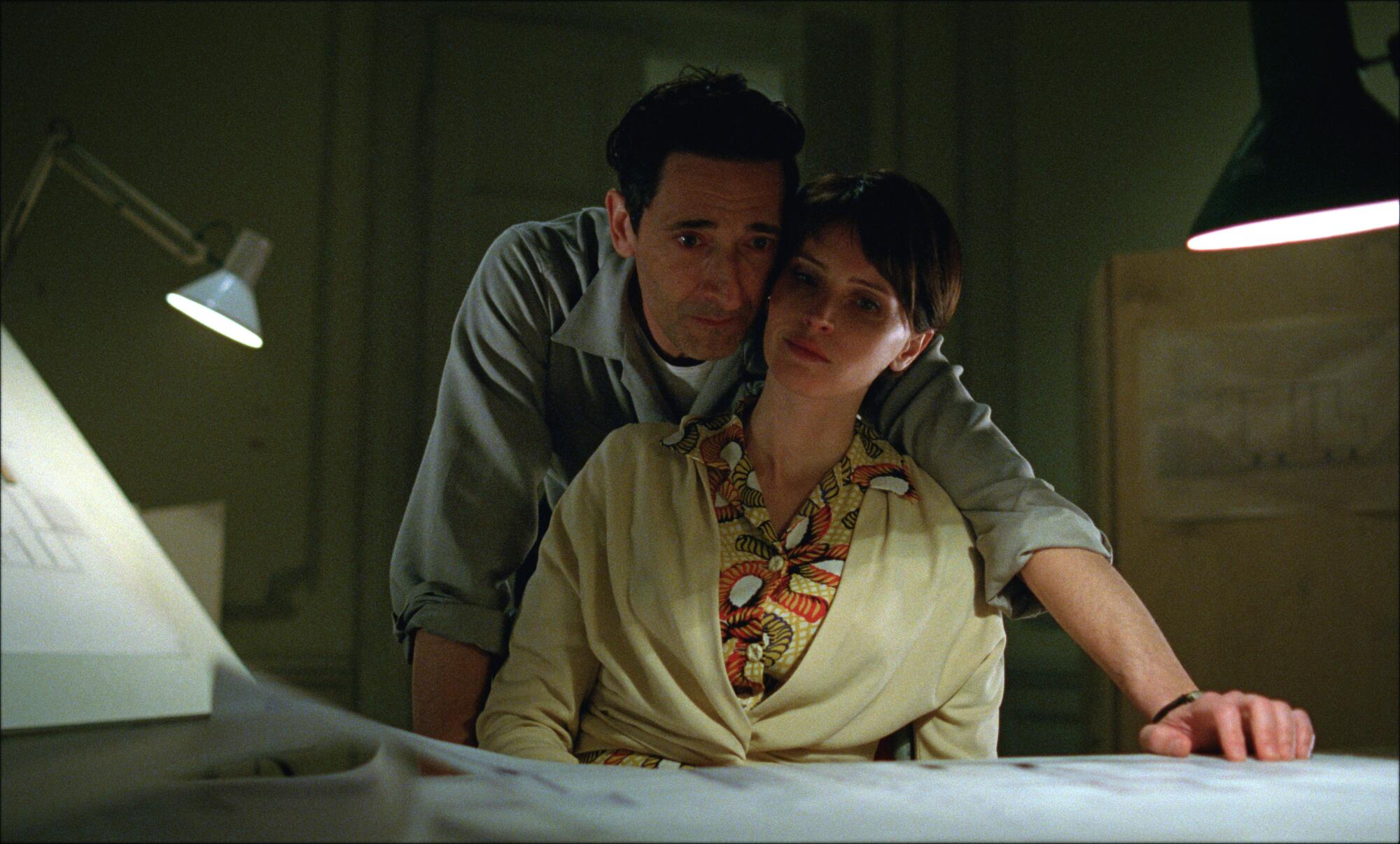 A man leans over a woman sitting at a desk looking at papers in "The Brutalist."