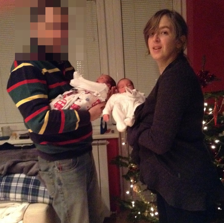 A woman and a man hold twin babies near a Christmas tree.  The man's face is blurred.