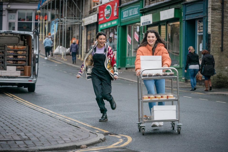 Two women hurrying down a street, pushing a cart of baked goods for a charity fete.