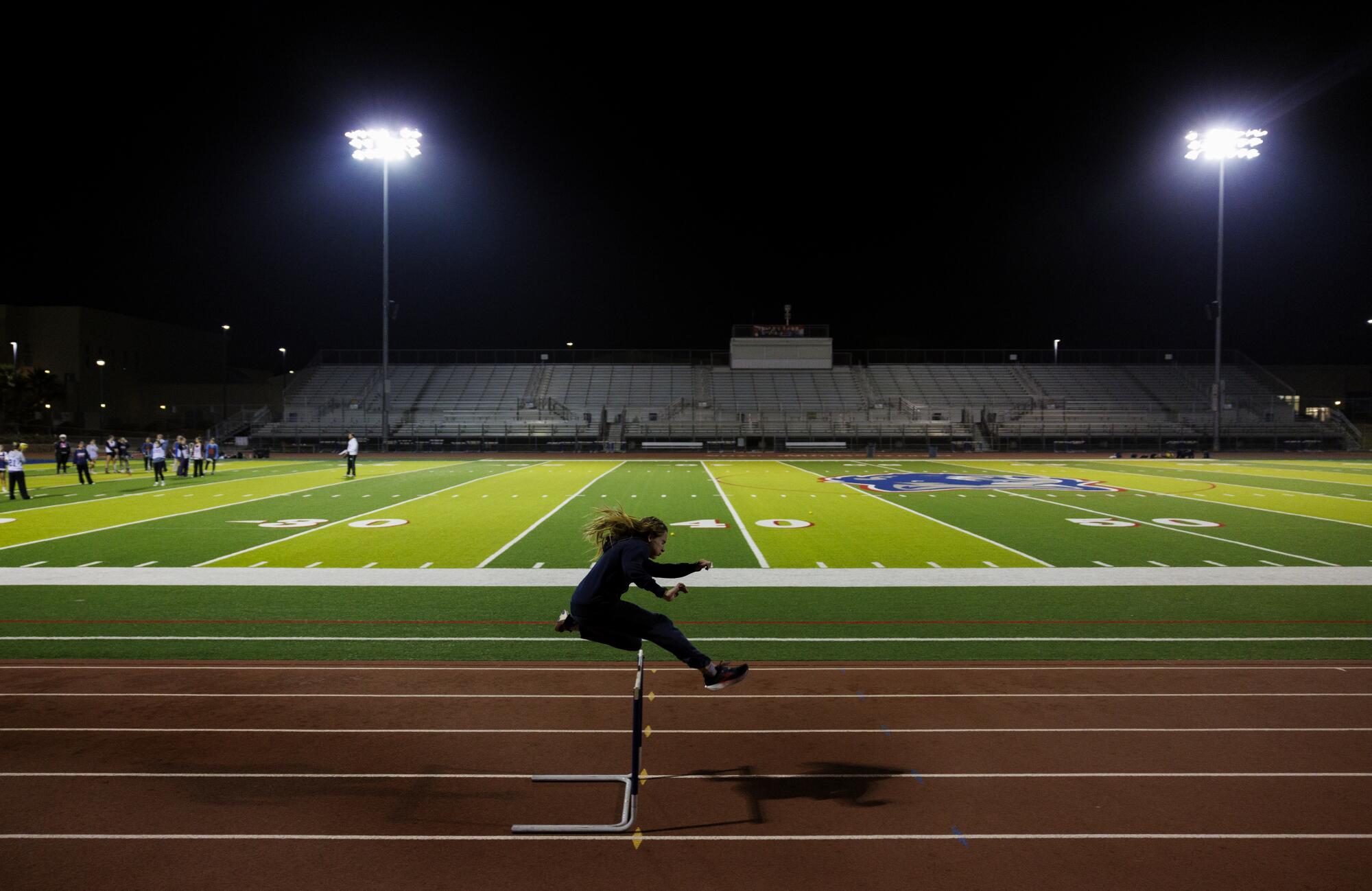 A young athlete clears a hurdle on a track at night. 