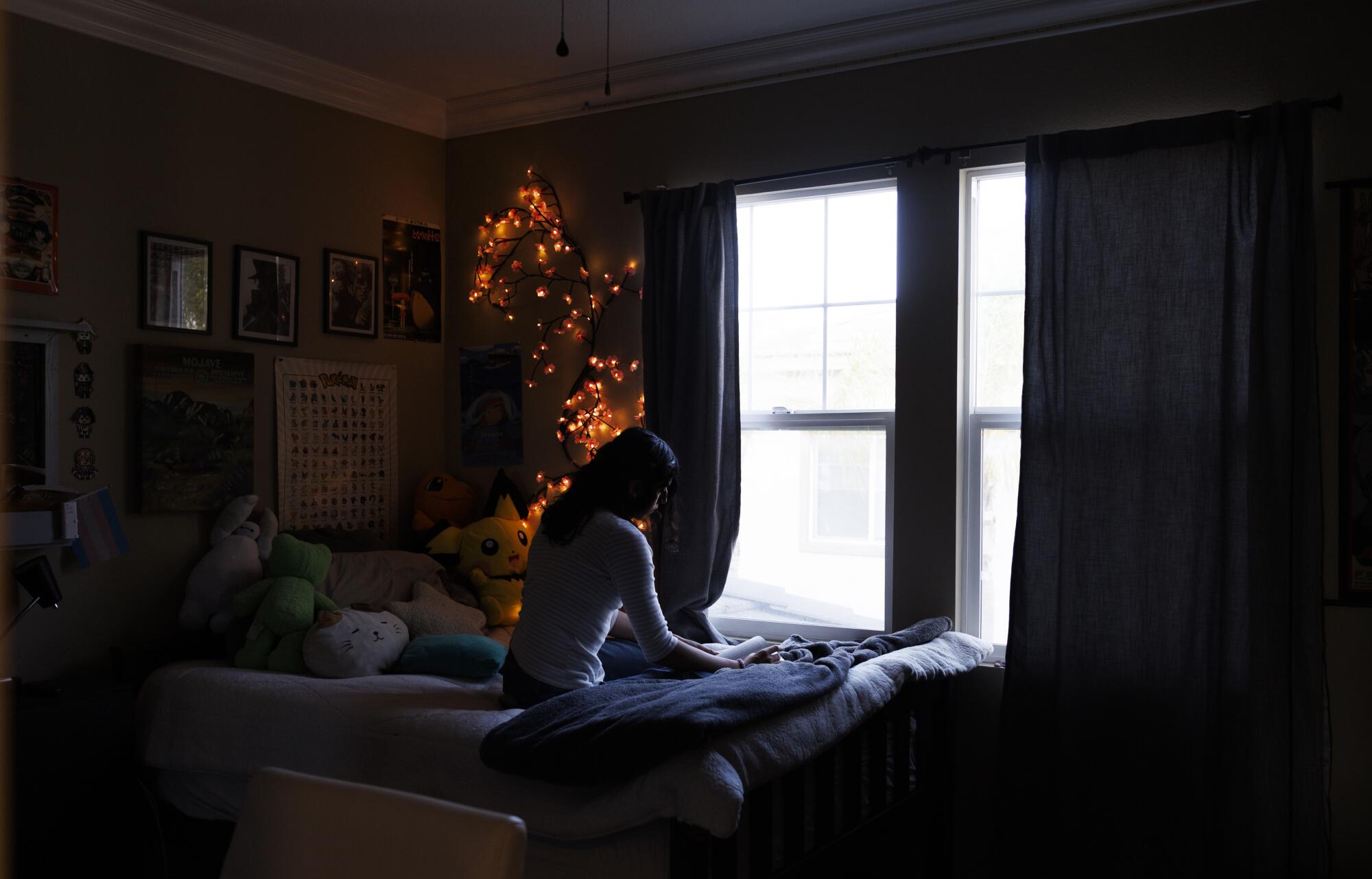 A teenager seated on her bed gazes out a window. 