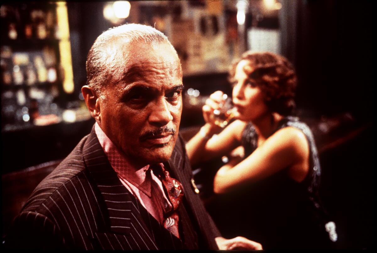 A man at a bar stares into the lens. A woman sits behind him at the bar, drinking.