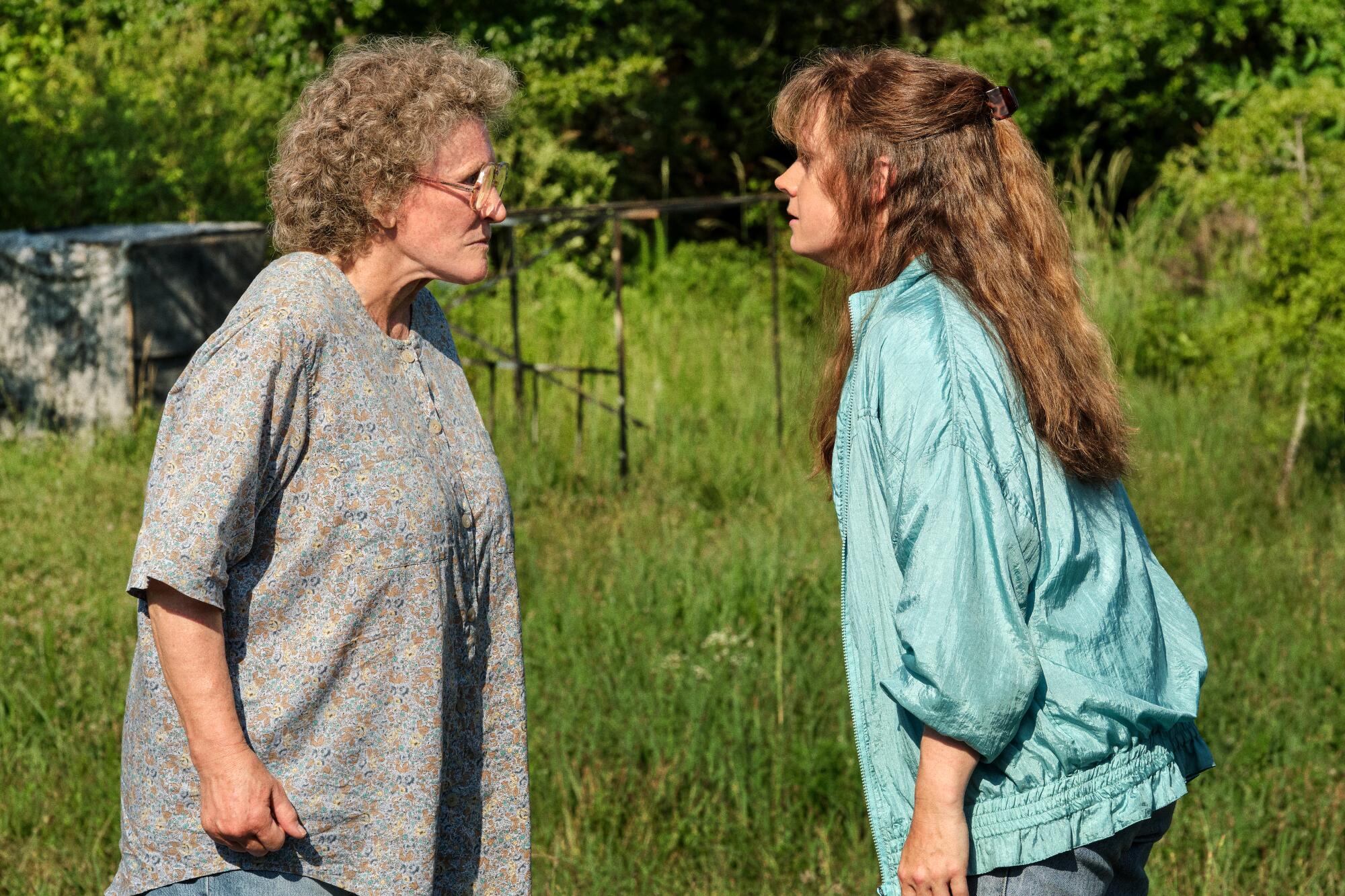 Two women, one older, one younger, stand facing each other in a field, looking angry and stubborn