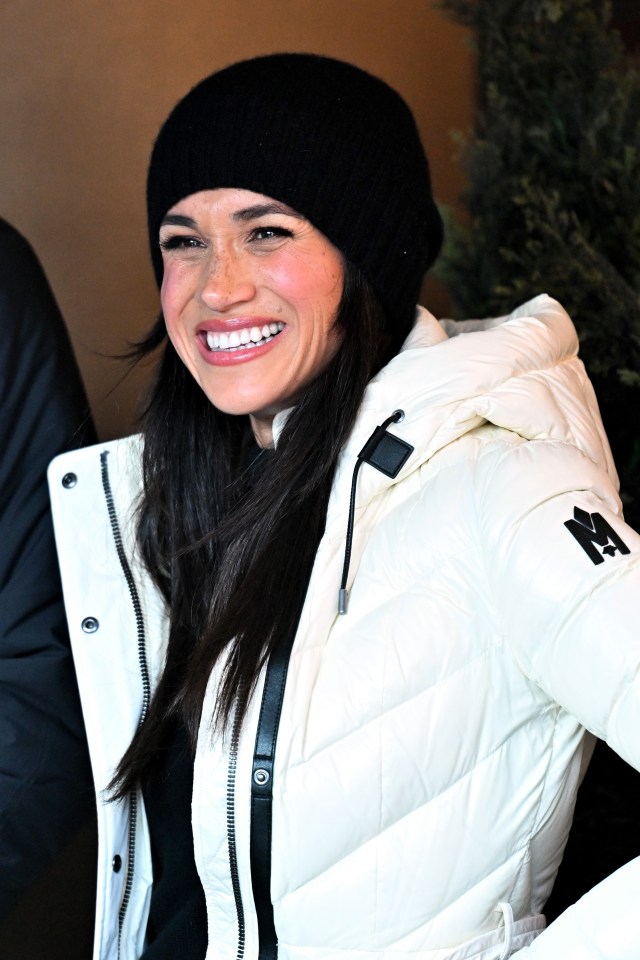 Meghan Markle smiling while wearing a white puffer jacket and black beanie.